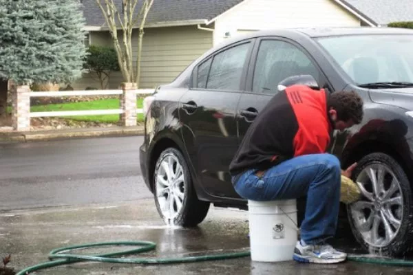 A picture of a guy washing his car on the driveway in the suburbs