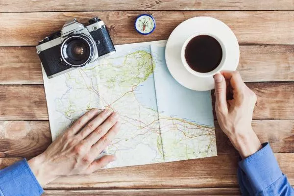 It’s very important to sit down, study the maps and make a travel plan in advance. two male hands, one holding a cup of tea and the other put on a map on a table where also places a camera and a compass